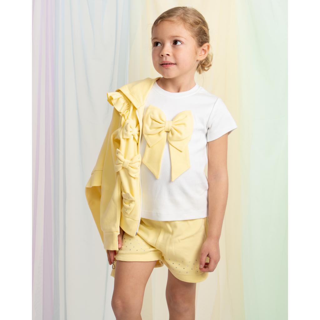 Model wearing a Caramelo yellow three‑piece short set for girls, featuring a bow T‑shirt, matching shorts and a ruffle‑trim jacket