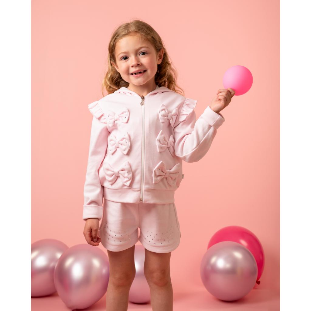 Model wearing a Caramelo pink three‑piece short set for girls, featuring a bow T‑shirt, matching shorts and a ruffle‑trim jacket