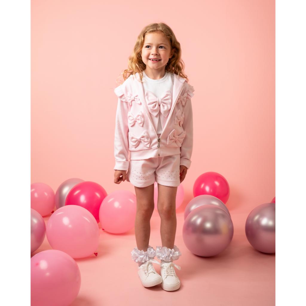Model wearing a Caramelo pink three‑piece short set for girls, featuring a bow T‑shirt, matching shorts and a ruffle‑trim jacket