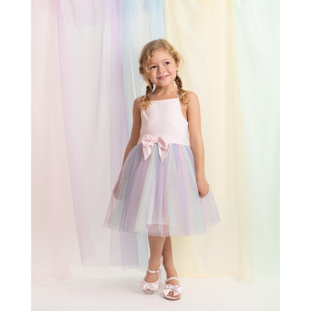 Caramelo rainbow pastel dress features a soft pink bodice with delicate straps and a statement bow at the waist, paired with a layered rainbow‑tulle skirt for a magical, floaty finish.