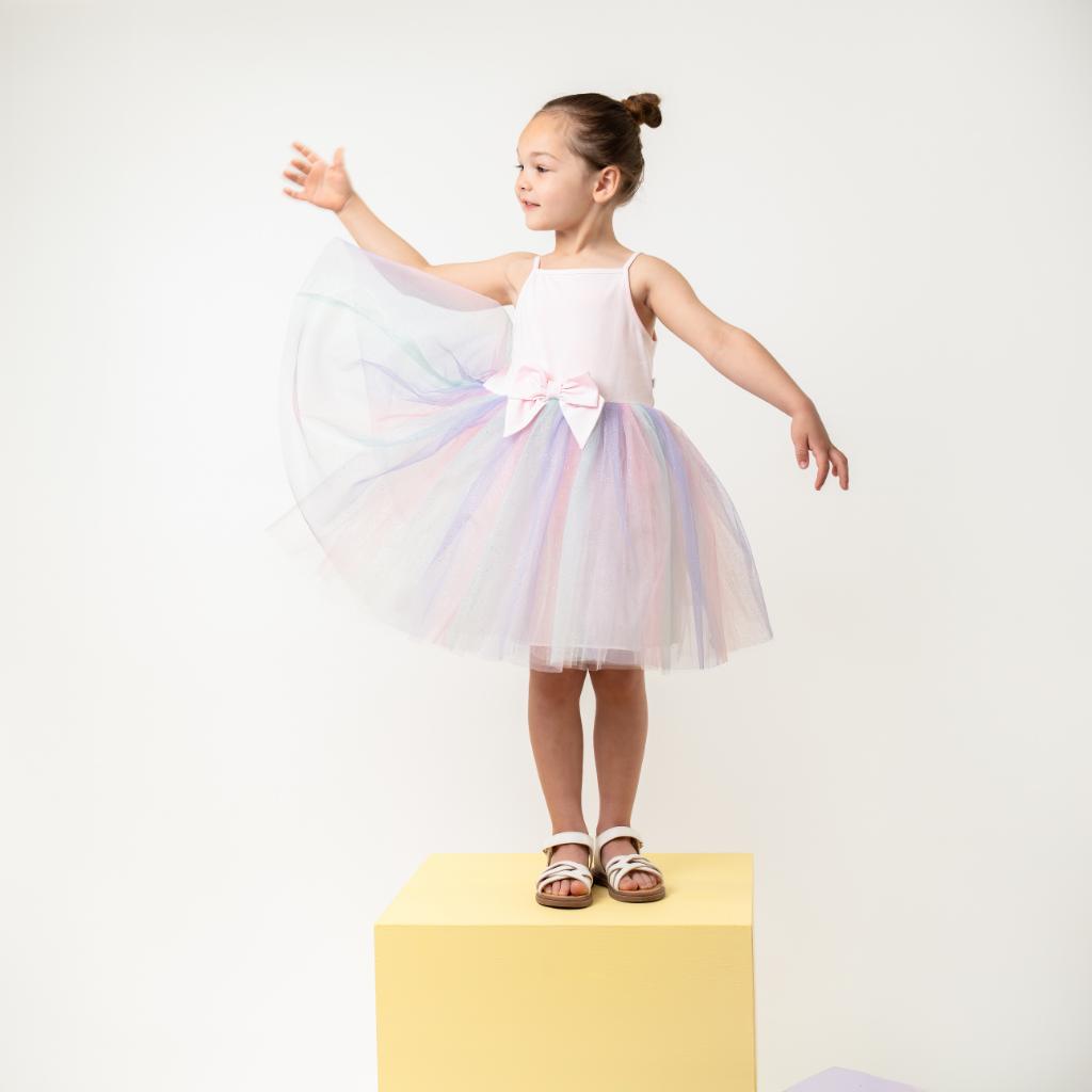 Model wearing a Caramelo rainbow pastel dress features a soft pink bodice with delicate straps and a statement bow at the waist, paired with a layered rainbow‑tulle skirt for a magical, floaty finish.
